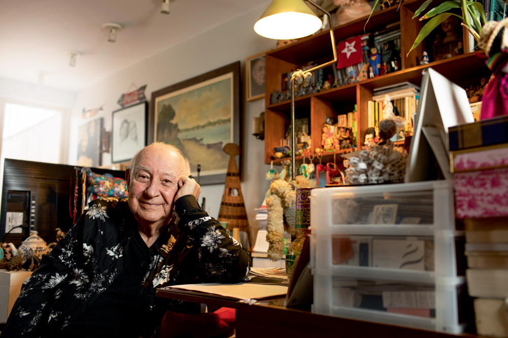 Sergio olha para a câmera, sorrindo sem mostrar os dentes, apoiando a cabeça na mão, que por sua vez está apoiada com o cotovelo na mesa. No entorno, é possível ver vários itens de sua sala, como quadros e livros.