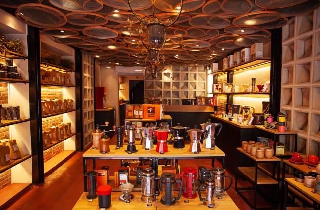 Achados Elo – Moka Clube Loja de café com prateleiras de madeira à esquerda e à direita cobertas por sacos pardos de café e xícaras de cerâmica. Ao centro, estante de madeira e estrutura metálica preta com cafeteiras italianas, moedores manuais de café e prensas francesas.
