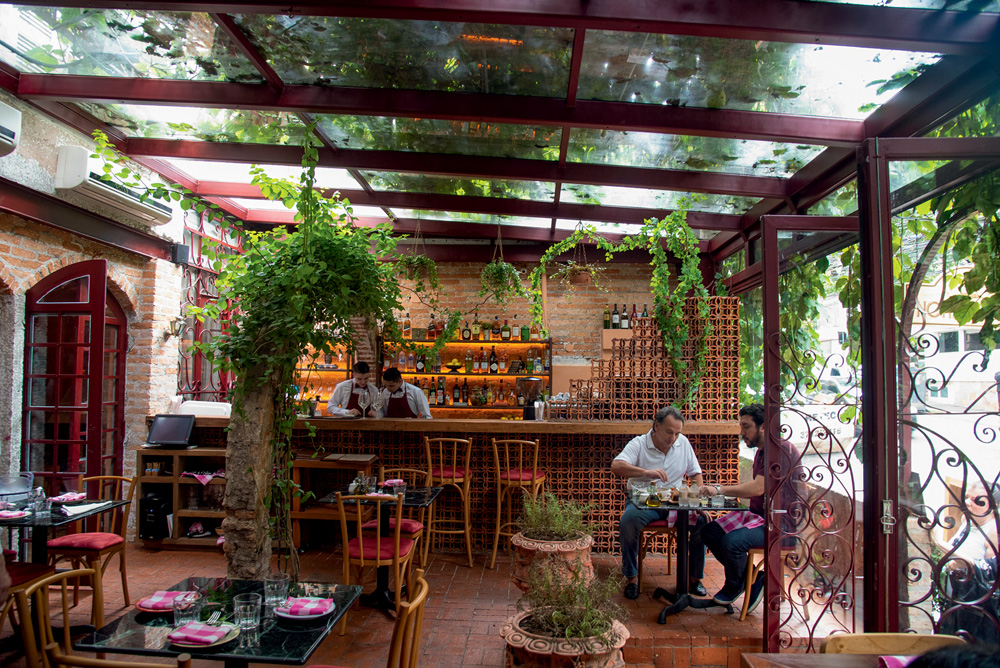 Espaço com plantas e duas pessoas sentadas em uma mesa. Ao fundo, dois homens trabalham.