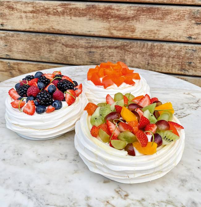 Confeitaria Dama – Dia das Mães Três pavlovas postadas em cima de uma mesa de granito, uma coberta de mix de frutas, outra de frutas vermelhas e a última, mais ao fundo, de caqui.