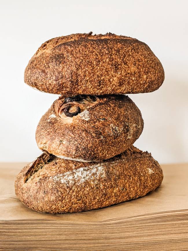 Trio de pães.jpg quatro pessoas agachadas com o rosto coberto por máscaras protetoras segurando canecas dentro de uma cozinha