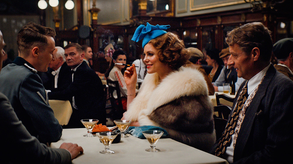 aespia.jpg Cena do filme A Espiã, em que a protagonista está em uma mesa de um restaurante com outros homens