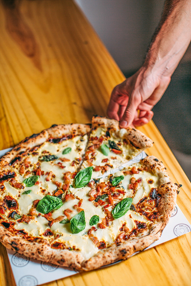 Mão retirando uma fatia da pizza que está sobre balcão de madeira