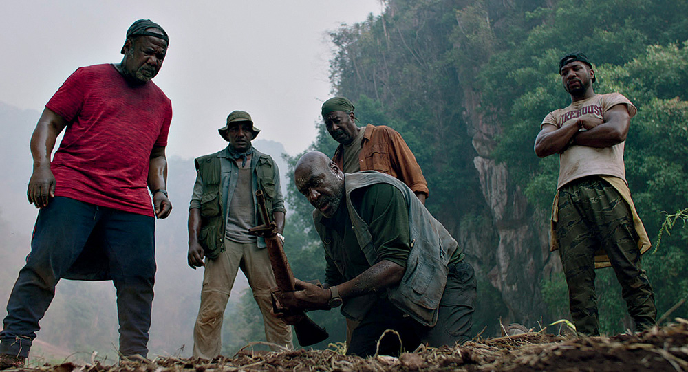 destacamento-blood.jpg Cinco homens negros olham para um objeto segurado por um deles perto do chão de terra