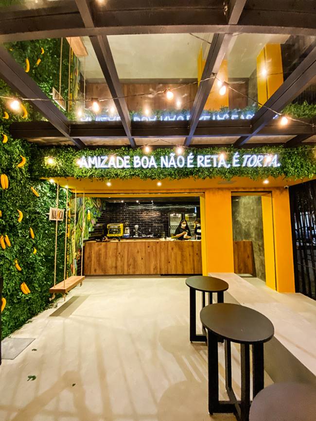 Nanica Salão da doceria Nanica com parede de plantas ao lado esquerdo, balcão ao fundo e banquinhos de madeira a direita