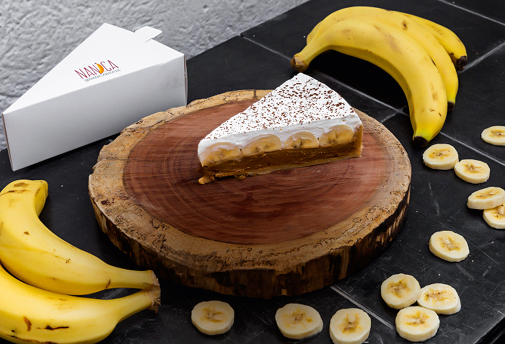 pedaço de torta banoffee em cima de tábua de madeira ao centro com parte de cacho de banana nas pontas inferior esquerda e superior direita; rodelas de banana por toda a lateral direita inferior