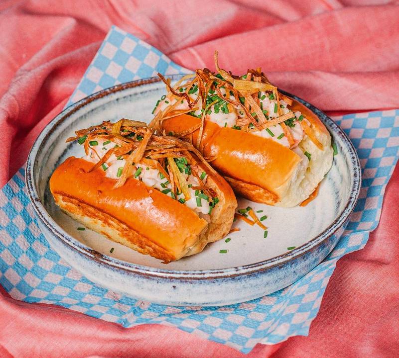 dois pequenos sanduíches em pão míni de cachorro-quente sobre prato em cima de guardanapo quadriculado azul e branco e tecido rosa