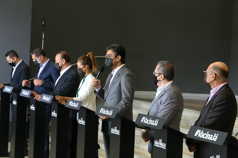 Foto lateral da coletiva de imprensa no Palácio de Bandeirantes. Em púlpitos distanciados, membros do governo do estado de São Paulo, todos de máscara, estão de frente para a imprensa.