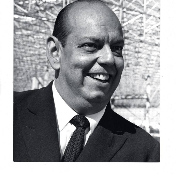 A imagem é um retrato, 3x4, do rosto de Caio. Ele está de terno e sorrindo.