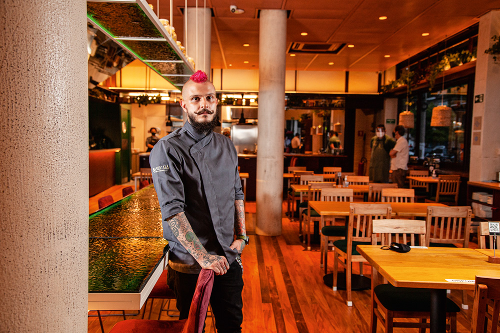 Chef Rafael Lorenti à esquerda, de dólmã e cabelo rosa, no salão do restaurante Basilicata Trattoria. Mesas à direita.