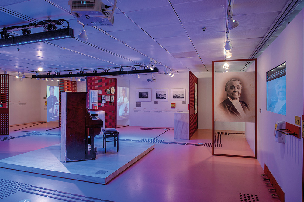 Crétidos_André Seiti (2).jpg Sala no Itaú Cultural. A luz é roxa e há pinturas, telões e gravuras que remetem a Chiquinha Gonzaga