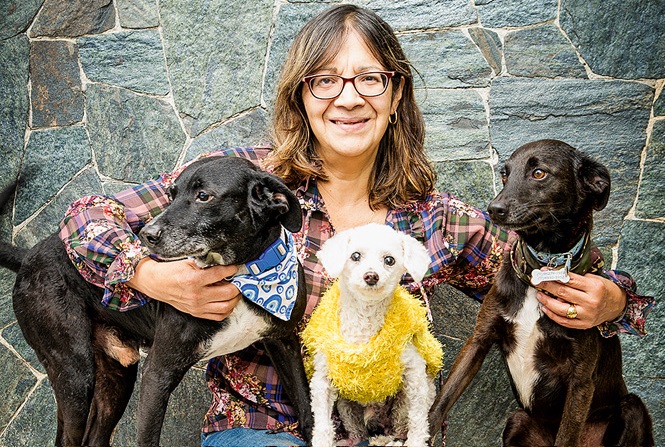 Uma mulher posa sentada em um banco com três cachorros, dois pretos e um branco