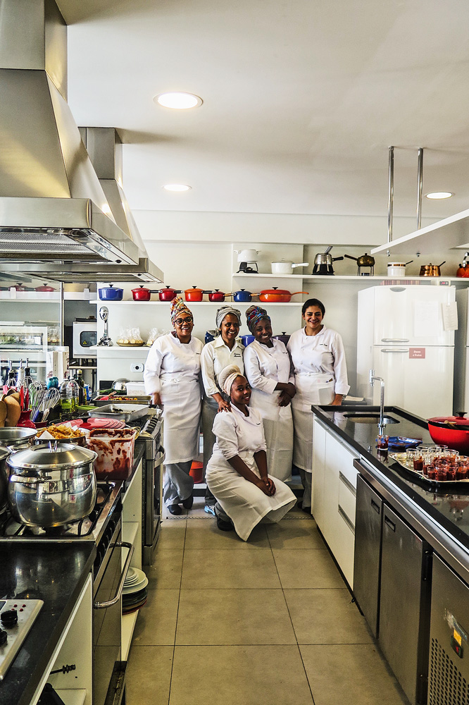 projeto-gastronomia-periférica-Formandas-turma-de-2019– GP Na cozinha ao fundo, aparecem cinco mulheres com uniformes de chef. Quatro estão de pé e uma ajoelhada para a foto. Todas aparecem sorrindo para a foto.