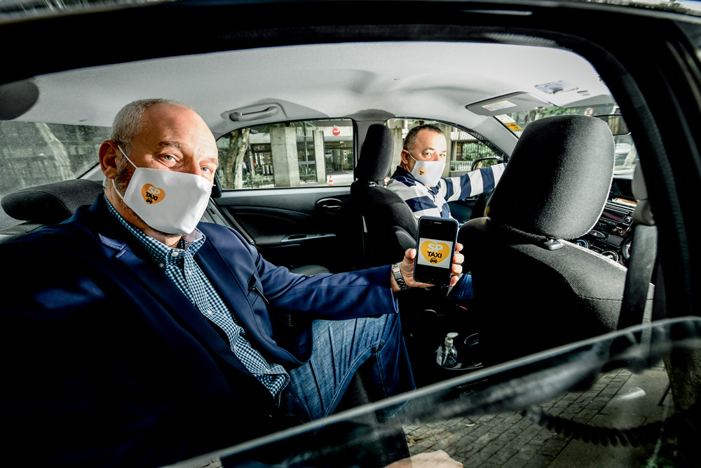 dois homens dentro de um carro, ambos olhando para a foto, um deles no banco do motorista e outro no banco traseiro. o homem do banco de trás, kasprik, segura celular com o SP taxi aberto