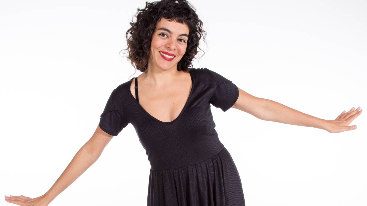 Mulher abre os braços e sorri olhando para câmera. Está de vestido preto em um fundo branco