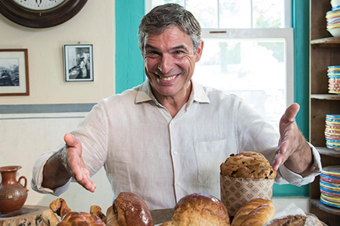 Olivier Anquier sorrindo com os braços meio abertos. Na parte inferior da imagem aparece um pouco dos pães e panetones de criação própria.