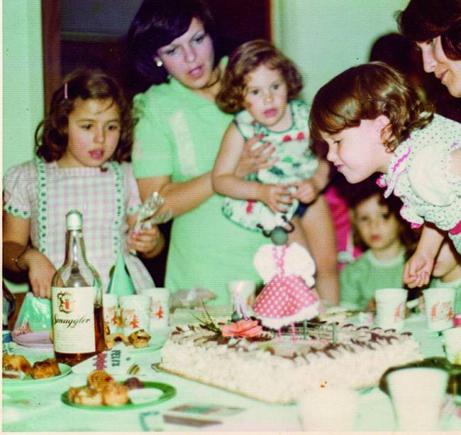 Família reunida ao redor de mesa com bolo de aniversário ao centro. Criança segurada por um adulto (à direita) sopra a vela do bolo.