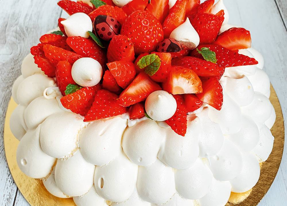 Imagem focada na pavlova, feita com merengue assado e chantili, em sua cobertura, existem vários pedaços de morangos cortados ao meio, enfeitando o doce.