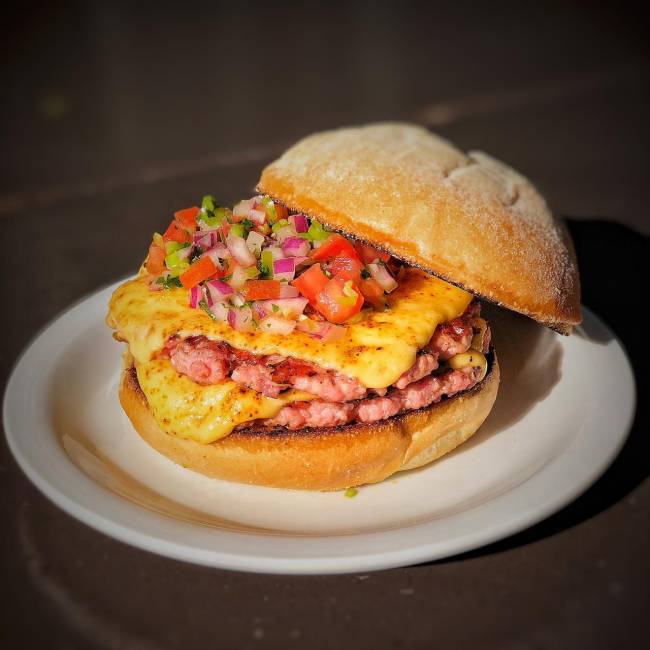 Dia do Hambúrguer – all_in Hambúrguer sobre prato de louça branca em cima de mesa de madeira. Dois discos suínos (rosados) com queijo e vinagrete por cima, com o pão superior afastado para o lado.