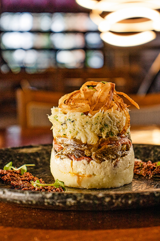 Prato de cerâmica sobre mesa de madeira com preparo "torre de bacalhau" em destaque: base circular de purê de batata sobreposta por conserva de pimentão, bacalhau desfiado e fatias de cebola frita.