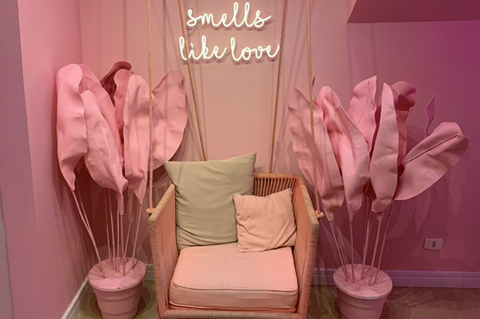 Espaço da cafeteria Café Cherie: poltrona com almofadas, vaso de flores falsas de ambos os lados e paredes cor de rosa.