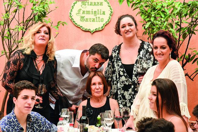 Speranza – Família Tarallo Família envolta de mesa de almoço com senhora matriarca ao centro e homem abaixado beijando o topo de sua cabeça.