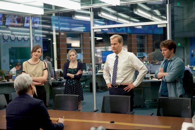 The-Newsroom A imagem apresenta personagens da série em uma sala de discussão, com uma mesa grande de madeira, em que há uma pessoa mais velha sentada em uma cadeira e outros quatro em pé em volta