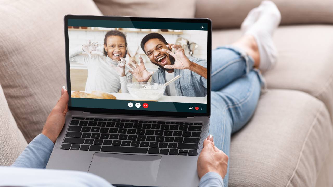 Pai e filha na tela do computador cozinhando. Vemos apenas os pés da mãe, com o computador no colo