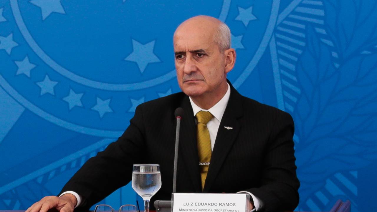 Ministro aparece usando terno, sentado em mesa durante coletiva de imprensa. Na sua frente, um microfone e um copo de água