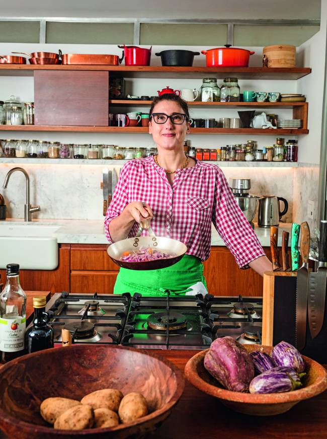 Paola Carosella vestida com camisa xadrez vermelho e branco segurando frigideira dentro de sua cozinha.