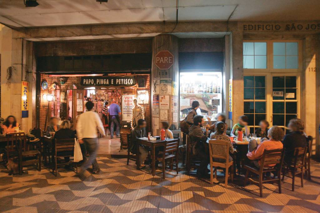 Fachada de bar com pessoas em pé e mesas na calçada