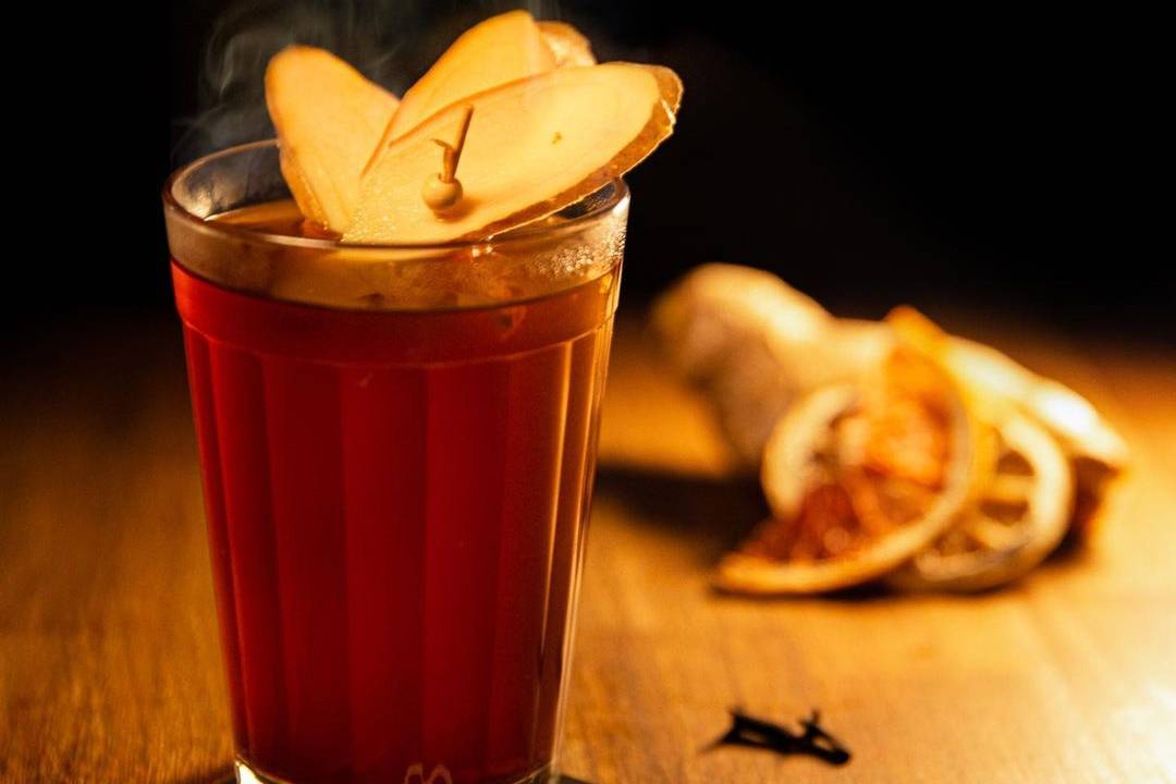 Copo americano com quentão fumegante coberto por fatias de gengibre sobre bancada de madeira.
