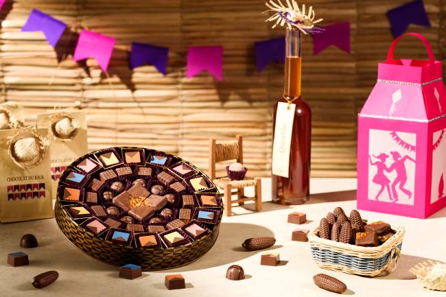 2021_05_27_chocolat-53 Caixa redonda de chocolate da Chocolat du Jour temático de festa junina ao lado de cestinha de cestinha com milhos de chocolate em cenário com banderolas rosas e roxas.