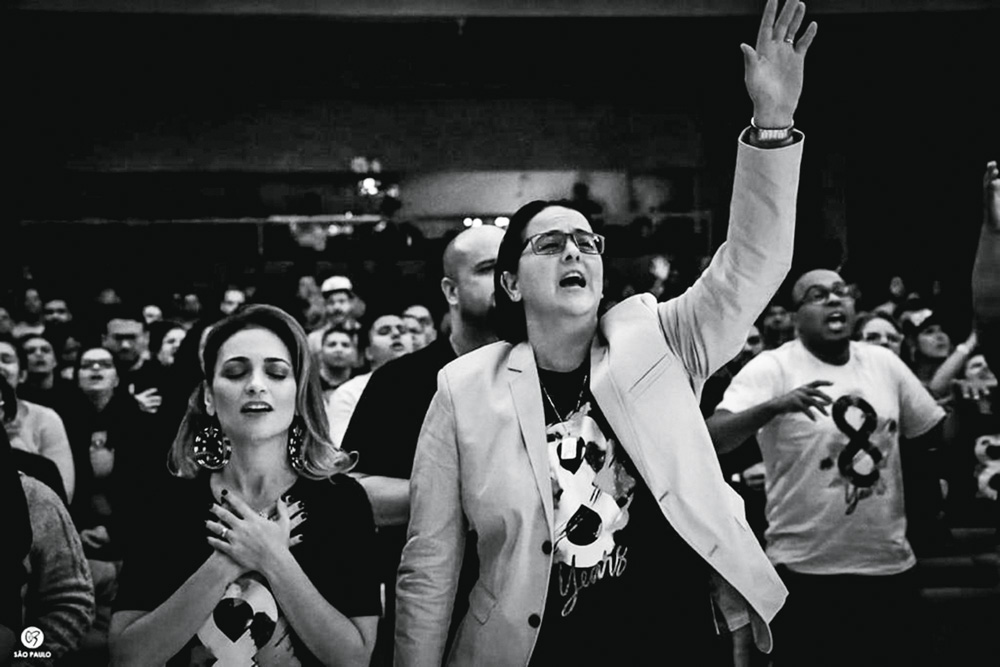 nosso-louco-amor-igreja-inclusiva-culto Rosania (à esquerda) e Lanna durante culto. Rosania está de olhos fechados com as mãos entre o busto e o pescoço e Lanna está com um dos braços levantados