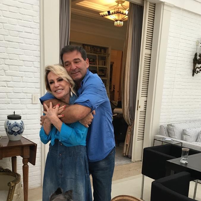 A imagem mostra Ana sendo abraçada por trás por Lucet. Ela coloca as mãos sobre os braços dele, ambos estão sorrindo.