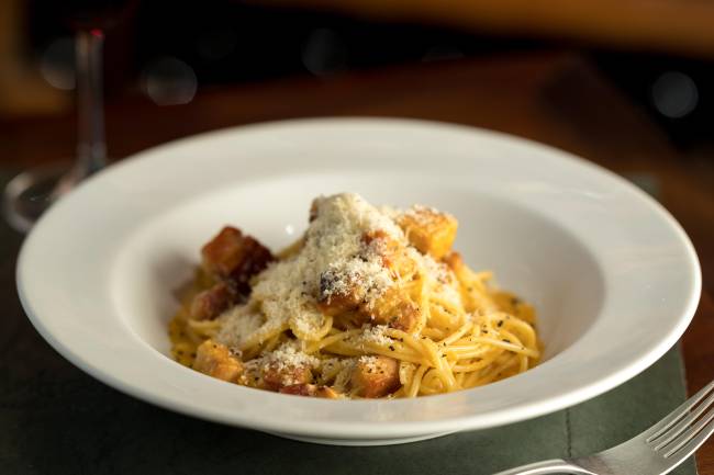 Carbonara de pato coberto por queijo em prato fundo de louça branca.