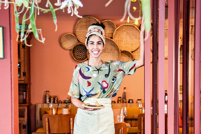 Bela Gil sorrindo na porta do restaurante Camélia Òdòdó enquanto segura um prato de palha.