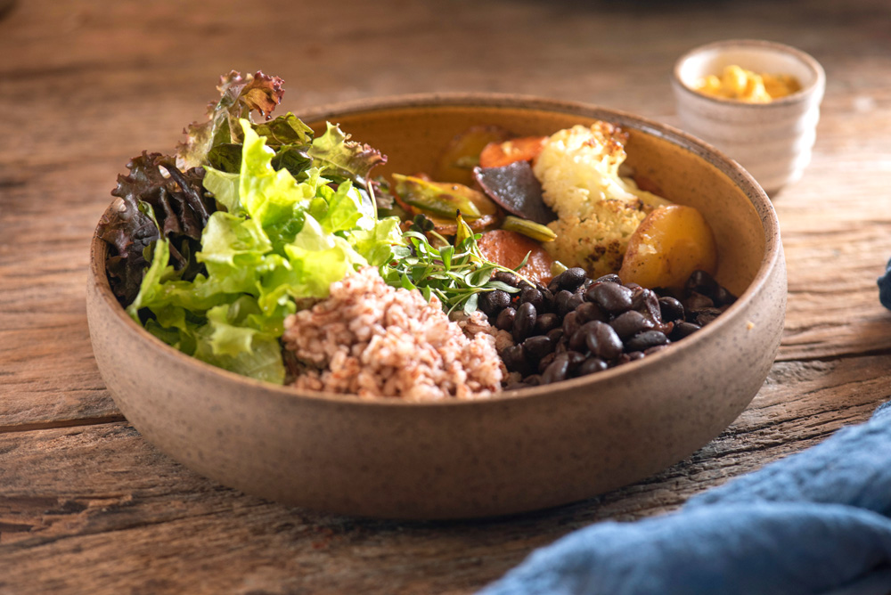 Camélia Òdòdó – Ocidente Prato de cerâmica sobre mesa de madeira. No prato tem arroz da terra (semipolido e trazido da Paraíba), feijão-carioquinha, vegetais salteados e salada verde mais farofa de castanhas