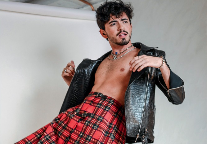 Foto mostra homem em um ensaio fotográfico. Ele está com uma perna levantada, jaqueta jeans, sem camisa e de saia quadriculada vermelha