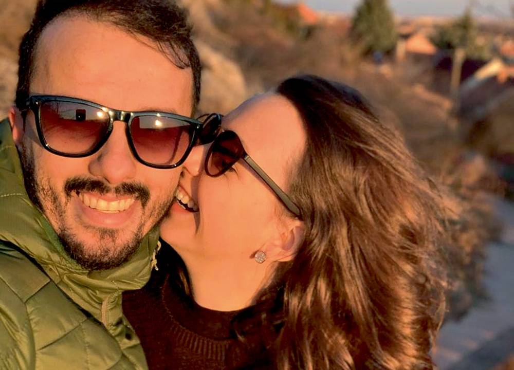 Roger e Raquel em selfie. Eles estão de óculos escuros com uma paisagem no fundo. Ele sorri para a foto enquanto ela está com o rosto colado na bocheca dele, com o nariz sendo amassado