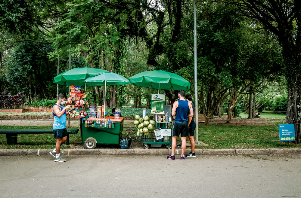 IBIRAPUERA ambulantes A imagem mostra um carrinho com guardo-sol e dois clientes olhando o que tem de opções nele dentro do Parque Ibirapuera