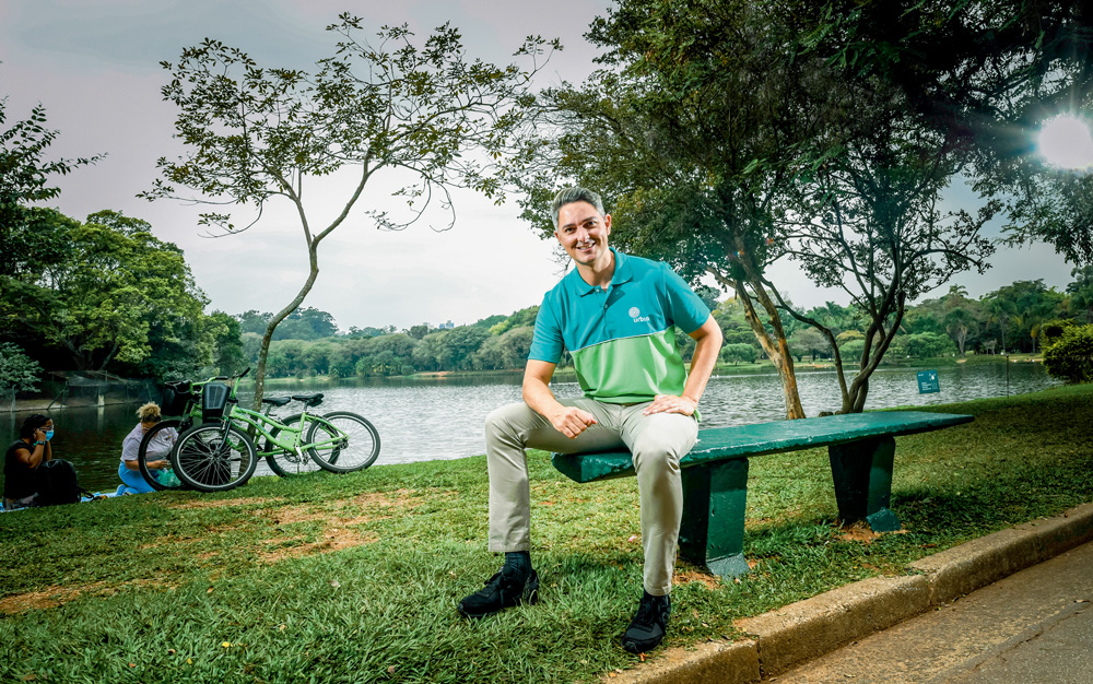 IBIRAPUERA_ lloyd A imagem mostra Lloyd sentado em um banco no parque Ibirapuera. Ele sorri para a câmera com a mão no seu joelho.