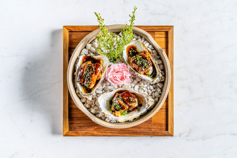 Makoto Em um objeto redondo com pedras para decorar, aparecem três ostras com foie gras. Uma flor de cor rosa aparece no meio como decoração.