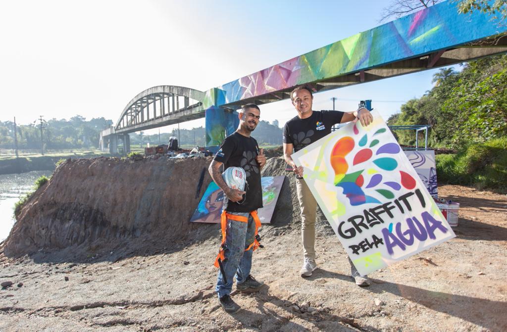 Projeto Graffiti Pela Água – Rodrigo Cordeiro e Gamão (LEO MARTINS) (1) Gamão (esquerda) e Rodrigo Cordeiro, idealizadores do projeto Graffiti pela Água. Ambos posam em frente a ponte colorida. Rodrigo segura placa com título