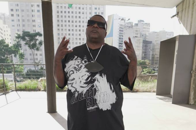 Alexandre Bane posa com mãos fazendo sinal de "hang loose" em galpão com fundo aberto. Veste camisa preta bem longa, óculos escuros e calça cinza.