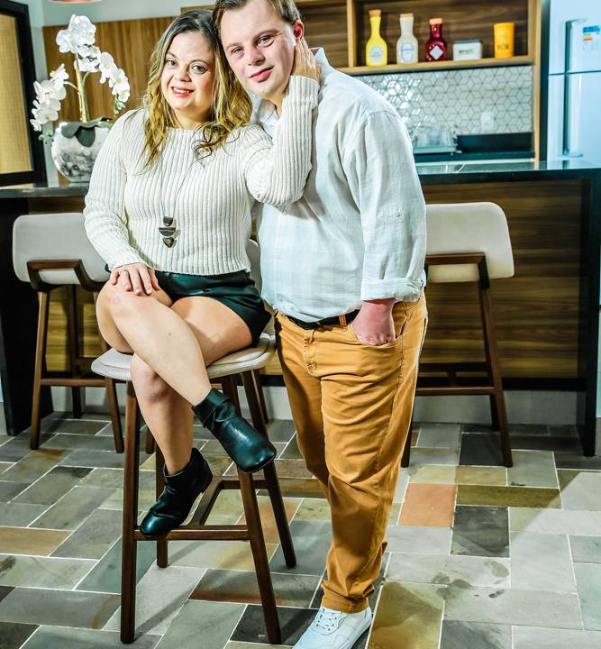 Tathi e Vinicius posando para a foto na sala de casa. ela está sentada em uma banqueta com seu braço esquerdo em volta do peito e pescoço de Vinicius, que está de pé do seu lado