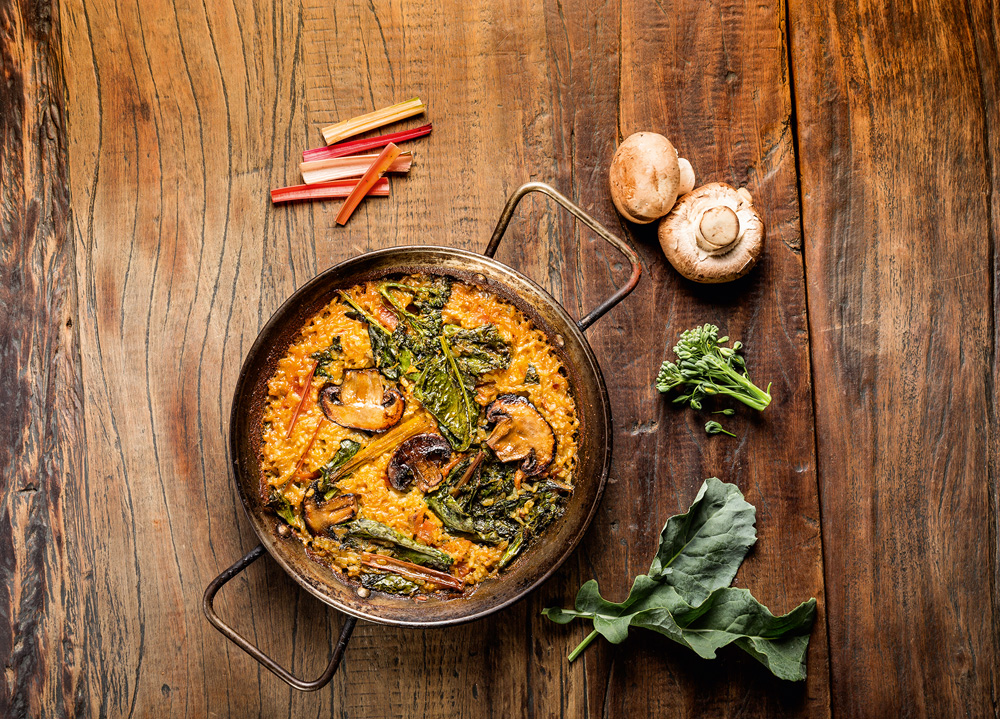 Sobre mesa de madeira, estão panela de cobre com arroz amarelado coberto por fatias de cogumelo tostadas e folhas verdes ao lado de itens frescos que fazem parte do preparo, como brocolis e cogumelos.