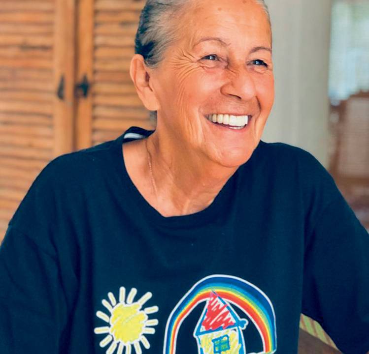 Tia Dag sorri sem olhar para a câmera. Cabelos grisalhos estão presos em um coque. Ela veste uma camiseta azul-marinho com logotipo da ONG Casa do Zezinho.