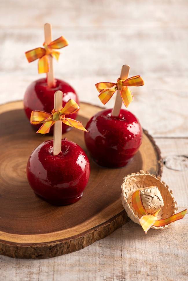 Casa Santa Luzia Três maçãs do amor (com caramelo vermelho por fora) postadas de cabeça para baixo com palito de madeira adornada por lacinho laranja sobre tábua redonda de madeira.