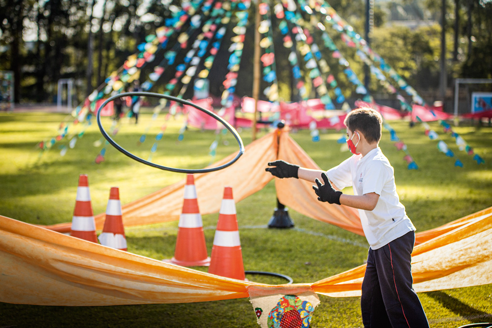brincadeiras juninas 2.jpg Uma criança joga um bambolê, que está no ar, em direção a quatro cones. Ao fundo, há bandeirinhas de festa junina
